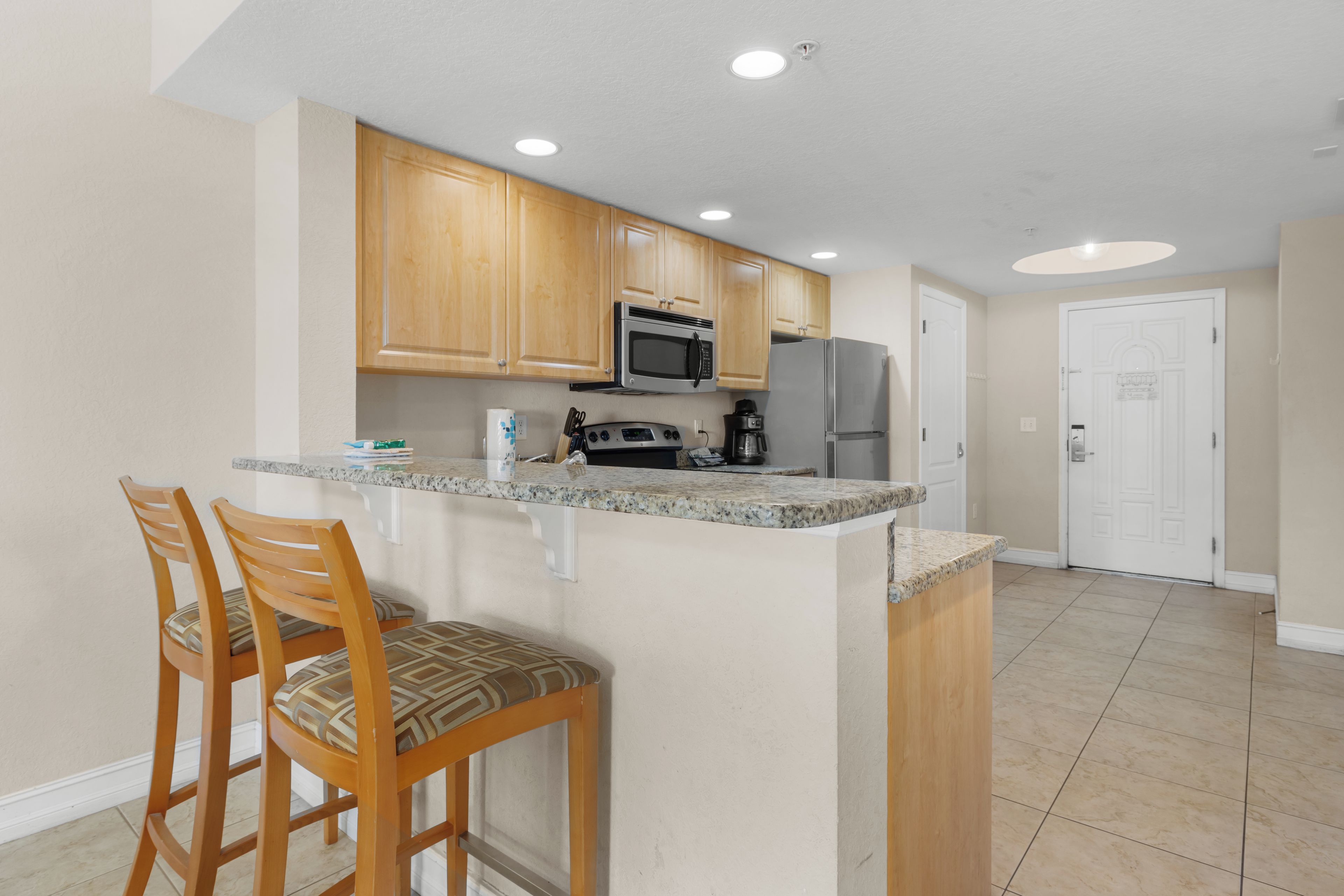 A kitchen with wooden cabinets, a microwave, a refrigerator, and a bar area with two stools.