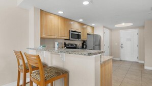 A kitchen with wooden cabinets, a granite countertop, a microwave, a refrigerator, and two bar stools. - Condo: 2BR/2BA by Beach, W/D, Pool & Parking (Treasure Island)