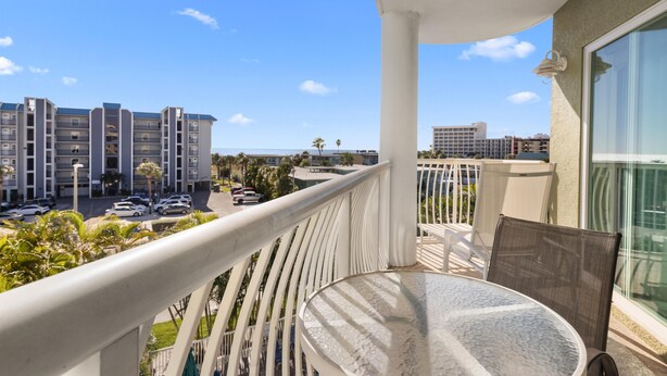 A balcony with a glass table and chairs, overlooking a parking area and buildings. - Condo: 2BR/2BA by Beach, W/D, Pool & Parking (Treasure Island)