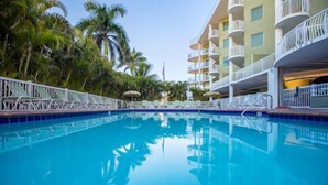 A swimming pool with a clear blue water surface, surrounded by palm trees and a multi-story hotel building with balconies. - Condo: 2BR/2BA by Beach, W/D, Pool & Parking (Treasure Island)