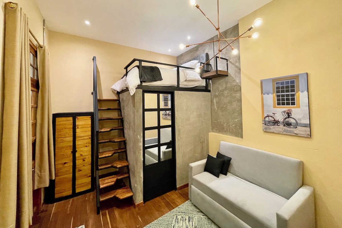 A compact room with a bunk bed, a staircase, a sofa, and a painting of a bicycle.