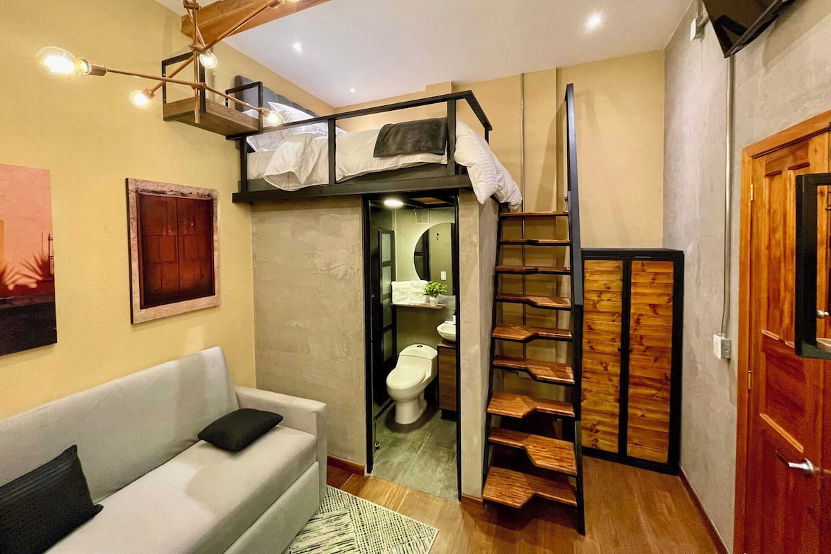 A compact living space with a bunk bed, a sofa, a wooden ladder, and a small bathroom.