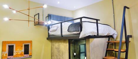 A dormitory room with bunk beds, a ladder, a wall-mounted shelf, and a ceiling light fixture.