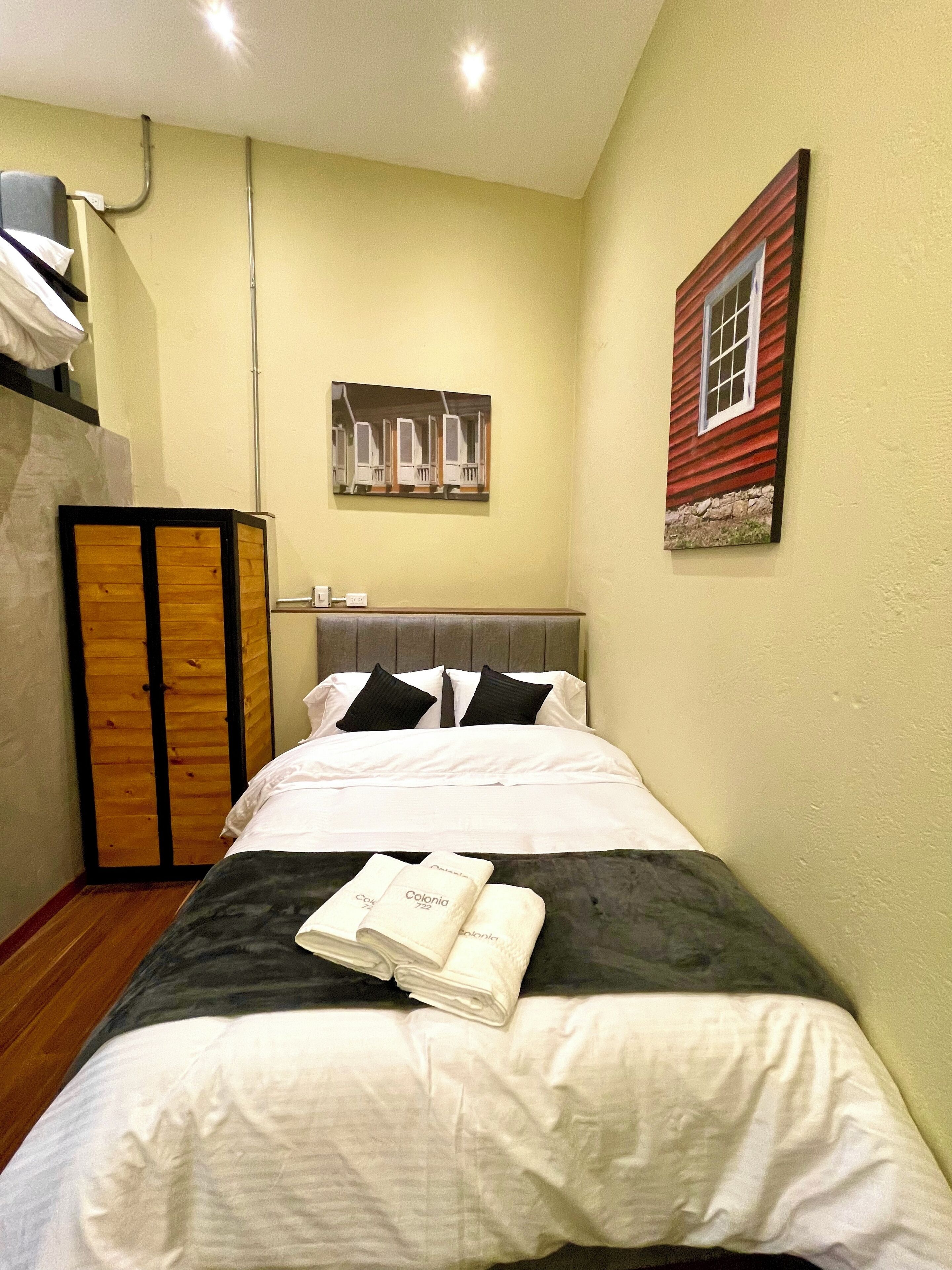 Una camera d'albergo con un letto, un armadio in legno, un quadro raffigurante una casa e una finestra con cornice rossa.