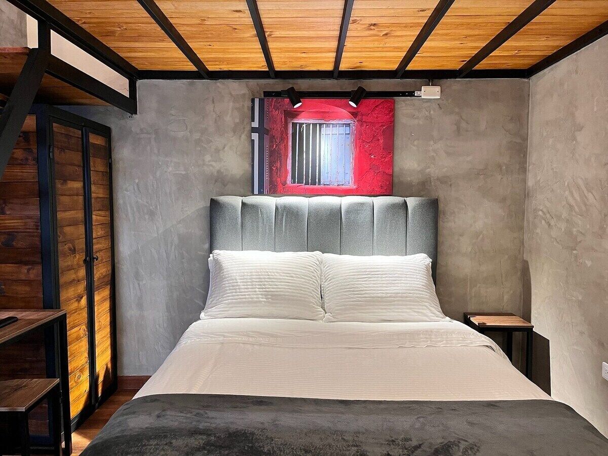 Un dormitorio con cama, cabecera, ventana con detalles rojos y techo de madera.