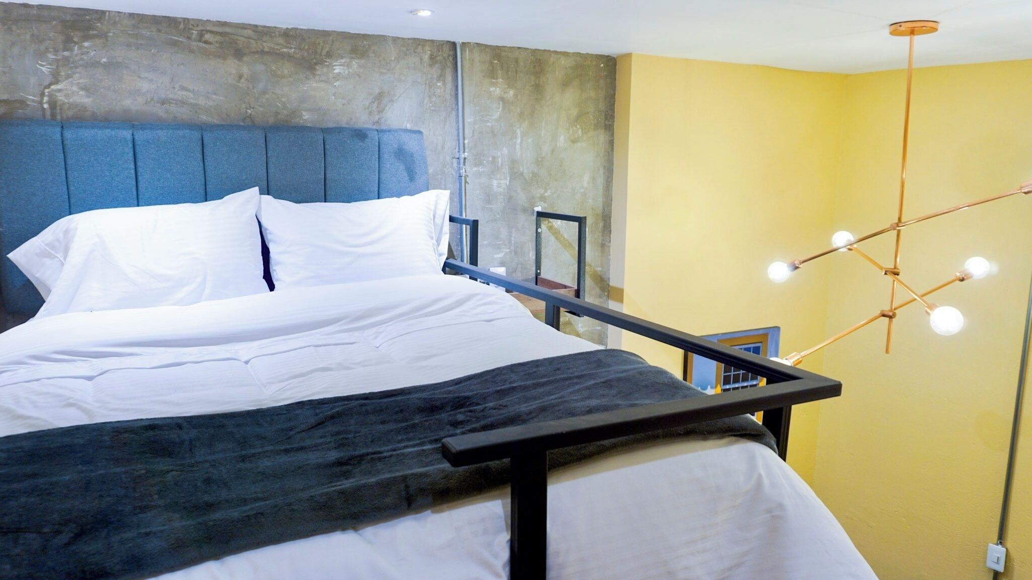 Quarto moderno com uma cama grande, cabeceira azul, luminária única e parede de destaque em concreto.