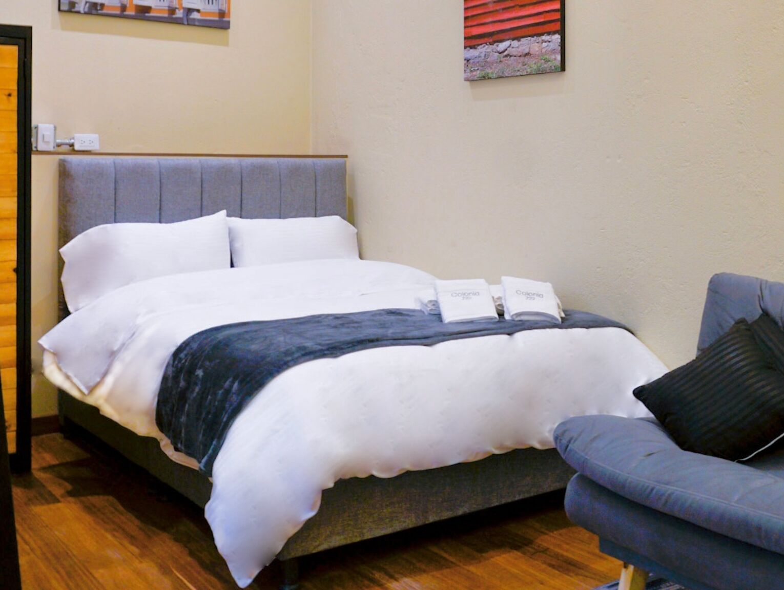 A neatly made bed with white bedding and a grey headboard, a grey sofa with a black pillow, and a small painting on the wall.