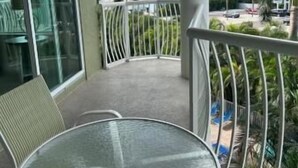 A balcony with a glass table and chairs, overlooking a beach and palm trees.