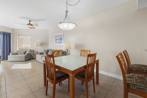 A dining area with a glass-top table surrounded by wooden chairs, a ceiling fan, and a pendant light. - Seaside 2BR Condo: Pool, Parking, & Beach (Treasure Island)