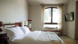 Ein Hotelzimmer mit einem großen Bett, einem Nachttisch mit Blumenvase, einem Fenster mit Vorhängen und einem an der Wand befestigten Fernseher.