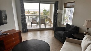 Un salon comprenant un canapé, une table basse, un téléviseur, un meuble en bois et un balcon offrant une vue sur la mer et les bâtiments.
