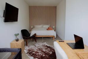 Un dormitorio con una cama, una silla, un escritorio con una computadora portátil, un televisor y una planta pequeña.