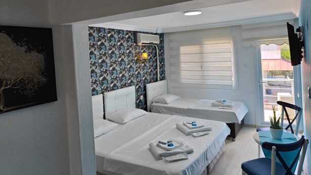 A hotel room with two beds, a wall-mounted air conditioner, a ceiling light, a window with blinds, a small table with a plant, and a chair. - Dafne Butik Pansiyon (Karaburun)