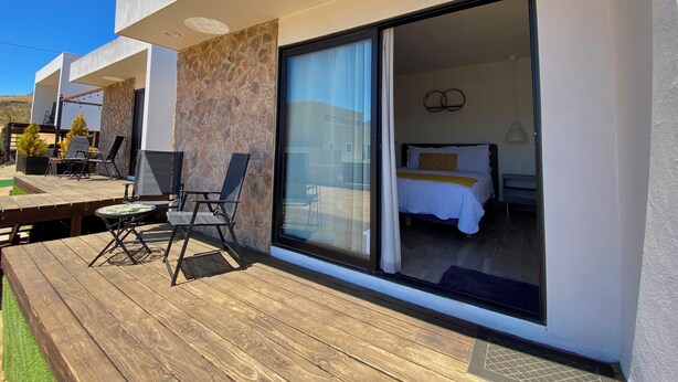 Una habitación de hotel moderna con un amplio ventanal corredero, una terraza de madera y una zona de estar con sillas.