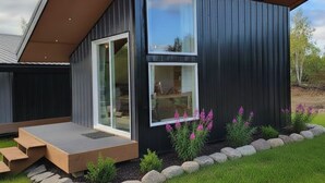 A modern house with a large glass door and windows, a wooden deck, and a garden with rocks and flowers. - Cozy Cabin on Private Airstrip (Houston)