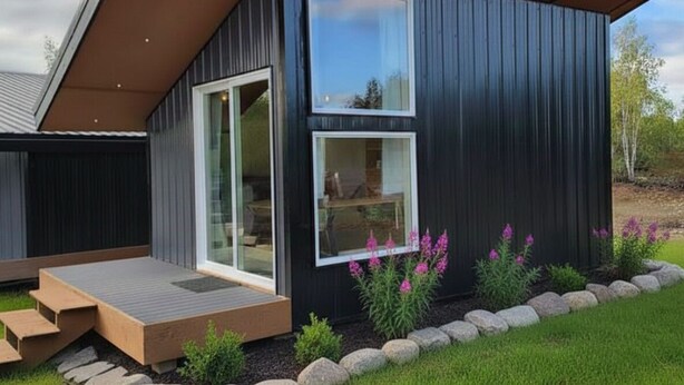 Ein modernes Haus mit großen Glasfenstern und einer Haustür, einer Holzterrasse und einem Garten mit Steinen und Blumen.