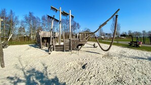 A playground with a wooden structure, including a swing set and climbing elements, set in a sandy area with trees in the background. - Familienfreundliches Domizil am Großen Meer mit Stangenofen. Haustierfreundlich! (Südbrookmerland)