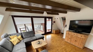 A modern living room with a grey sectional sofa, a wooden TV stand, and a balcony with a view. - Familienfreundliches Domizil am Großen Meer mit Stangenofen. Haustierfreundlich! (Südbrookmerland)