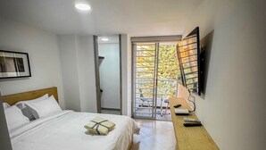 A hotel room with a bed, a wooden nightstand, a TV mounted on the wall, and a sliding glass door leading to a balcony with a view of trees. - Encantador Loft en Medellín (Medellin)