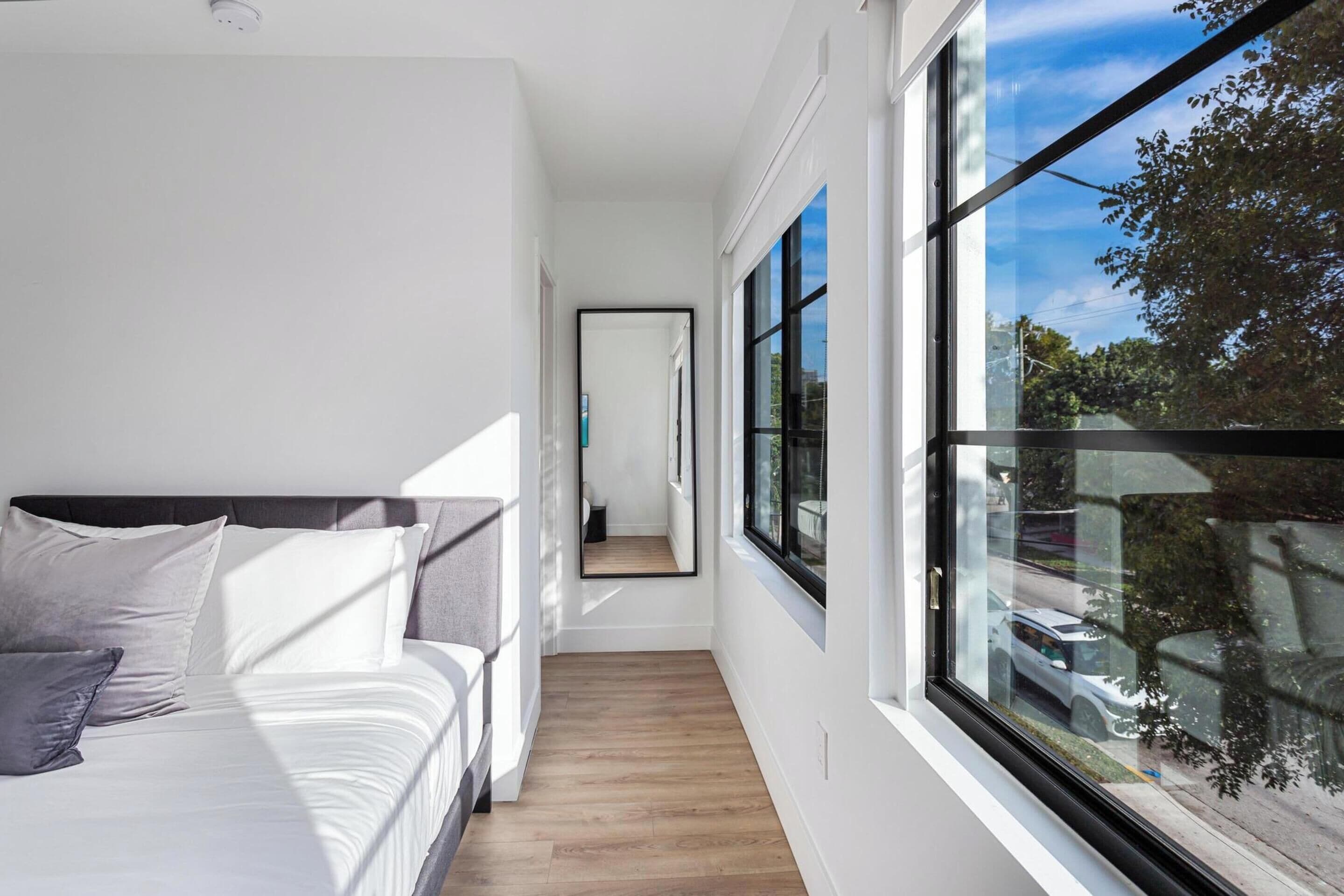 A modern bedroom with a large window, a bed with white linens, and a wooden floor.