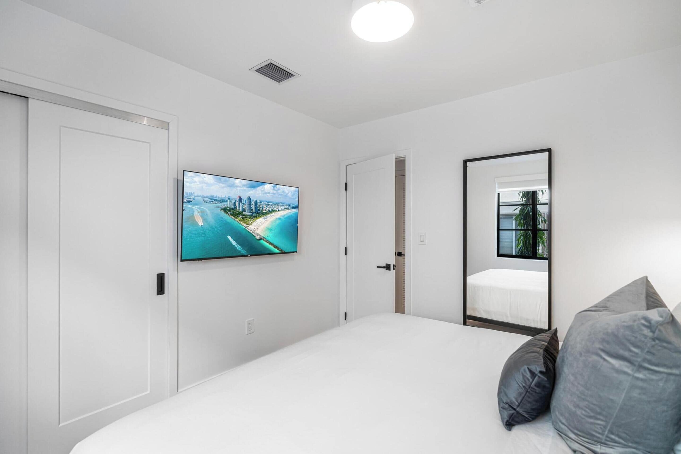 A bedroom with a large bed, a flat-screen TV mounted on the wall, a mirror, and a door leading to another room.
