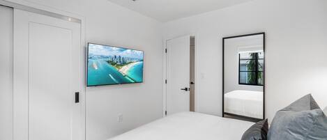 A bedroom with a large bed, a flat-screen TV mounted on the wall, a mirror, and a door leading to another room.
