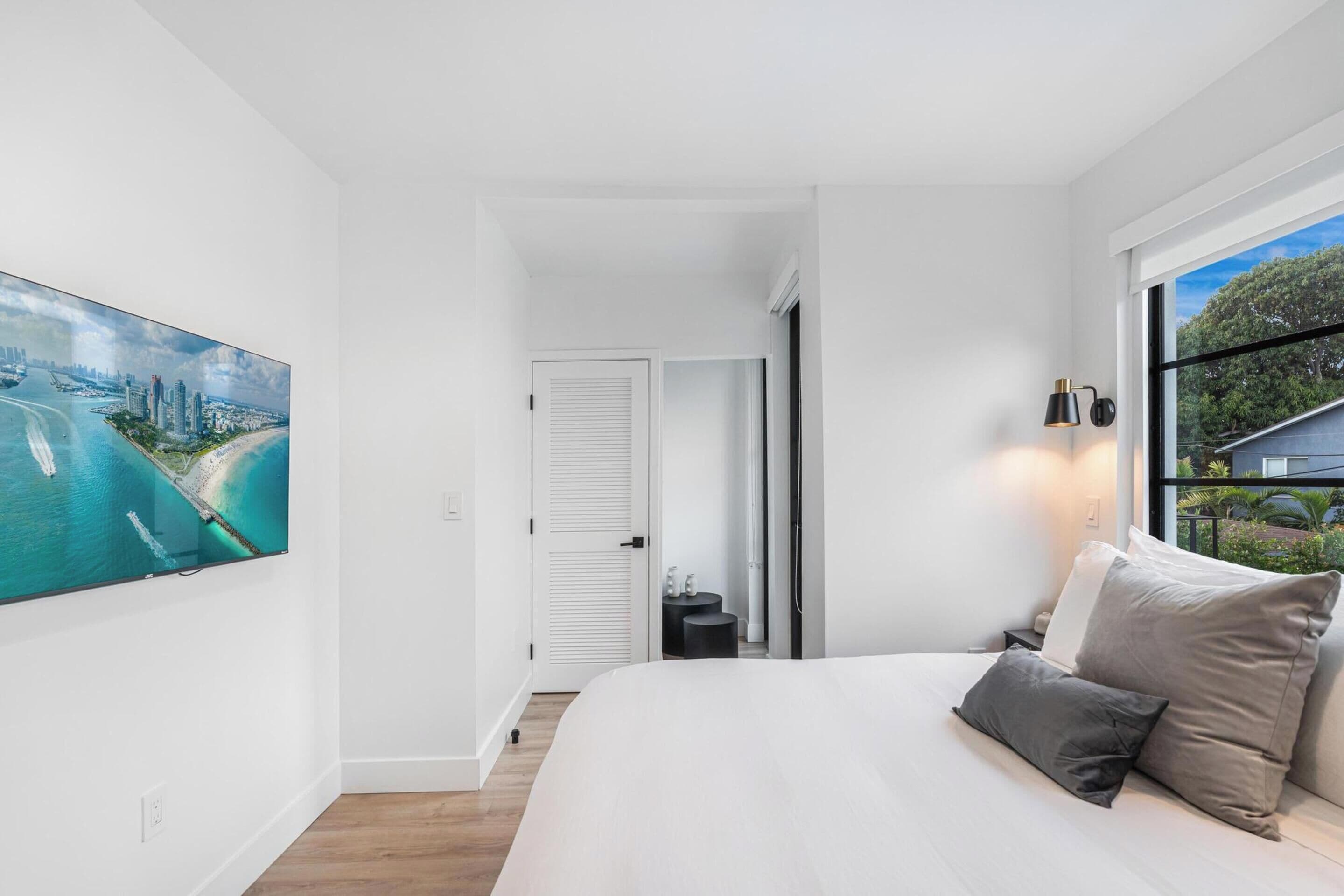 Ein modernes Schlafzimmer mit einem großen Bett, Blick auf eine Stadtlandschaft durch ein Fenster und einem Gemälde einer Küstenansicht.