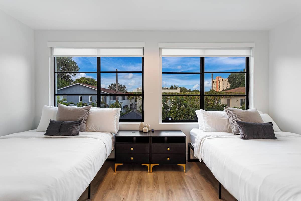 Ein modernes Schlafzimmer mit zwei Betten, einer schwarzen Kommode und großen Fenstern mit Blick auf die Stadt.