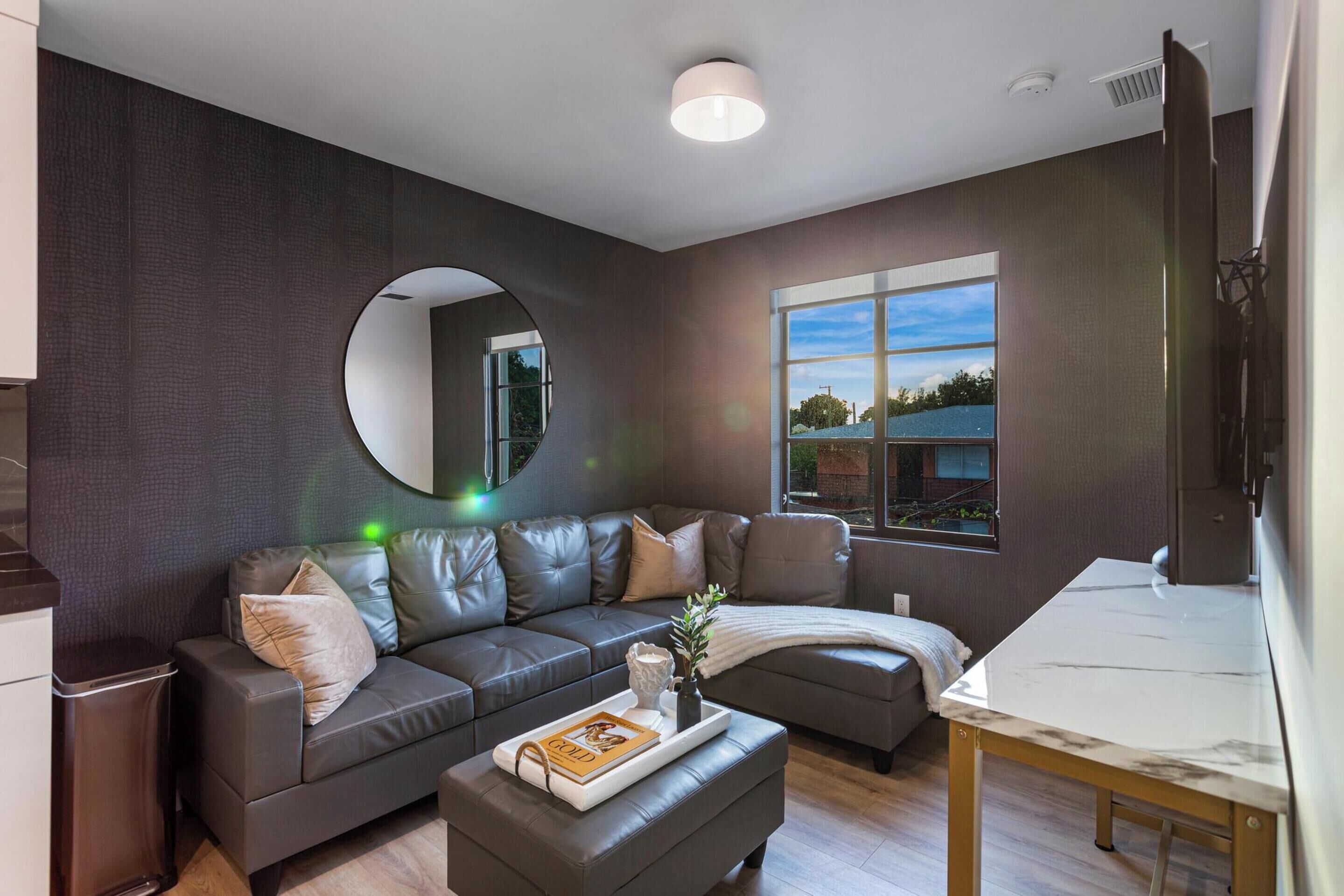 Una sala de estar moderna con una pared marrón oscuro, un espejo redondo, un sofá de cuero gris, una mesita con una bandeja y una ventana con vistas al exterior.