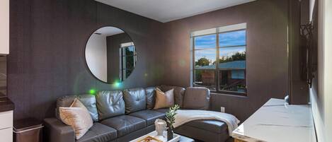 Una sala de estar moderna con una pared marrón oscuro, un espejo redondo, un sofá de cuero gris, una mesita con una bandeja y una ventana con vistas al exterior.