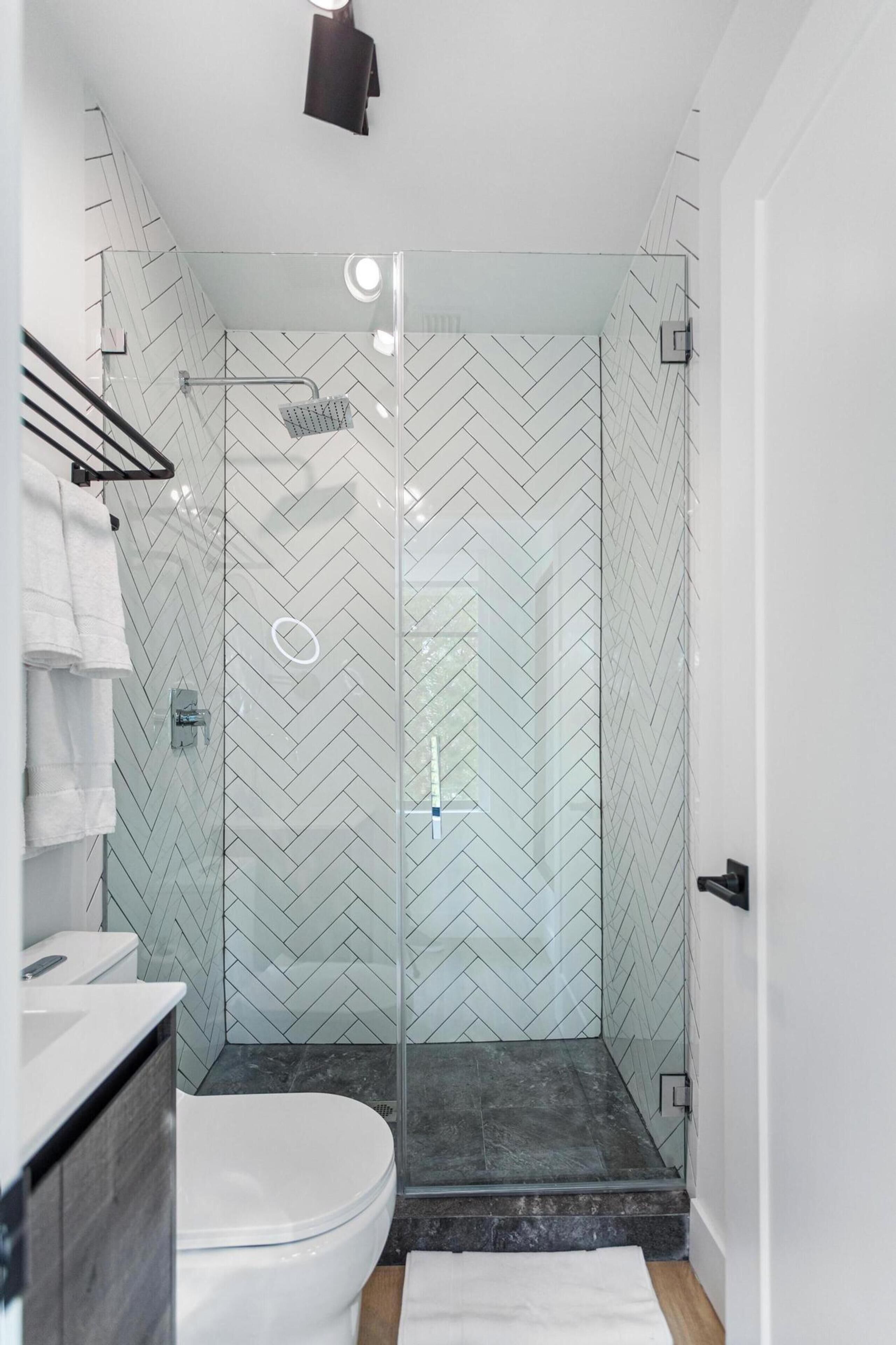 Une salle de bain moderne avec une douche à parois de verre, des carreaux blancs disposés en motif chevrons, des toilettes blanches et un plancher en bois.