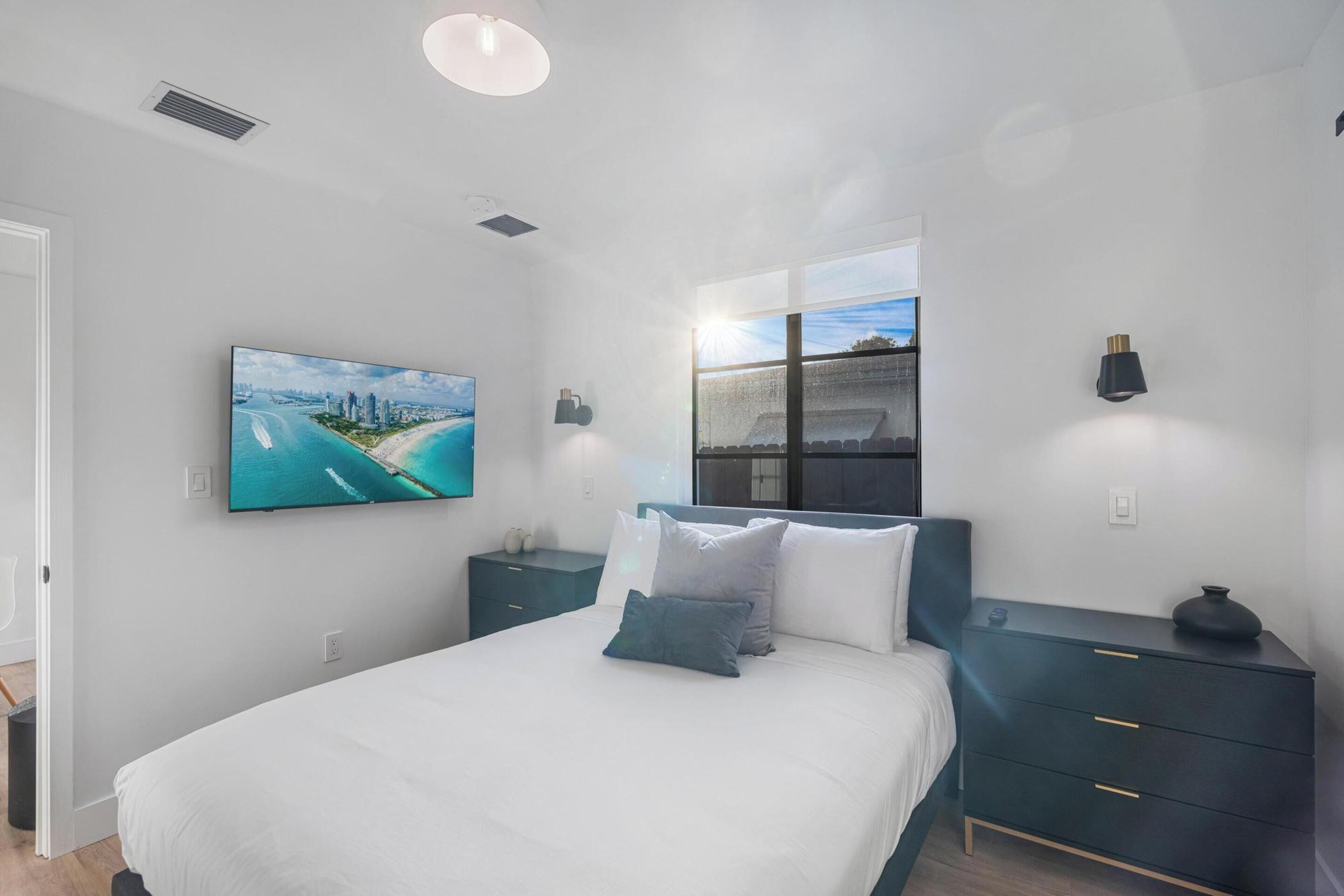 A bedroom with a bed, a nightstand, a wall-mounted TV, a window with a view, and a ceiling light.