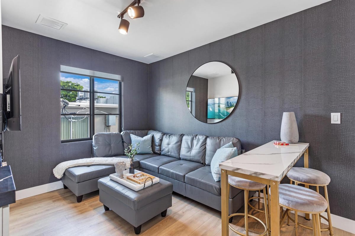 Ein modernes Wohnzimmer mit einem grauen Sofa, einem runden Spiegel, einem Esstisch mit Hockern und einem großen Fenster mit Blick ins Freie.