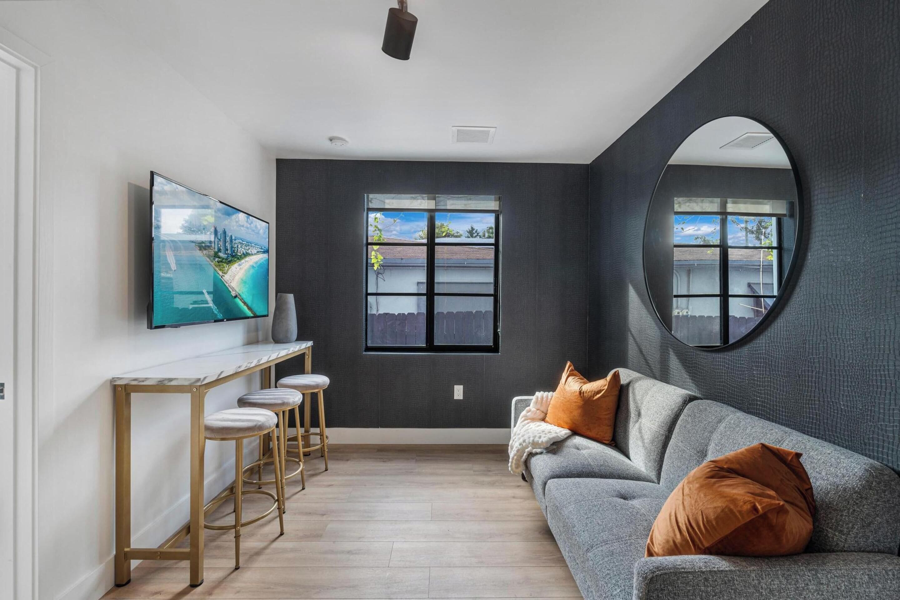 A modern living room with a grey sofa, a round mirror, a TV on a marble console, and a large window with a view of the outside.