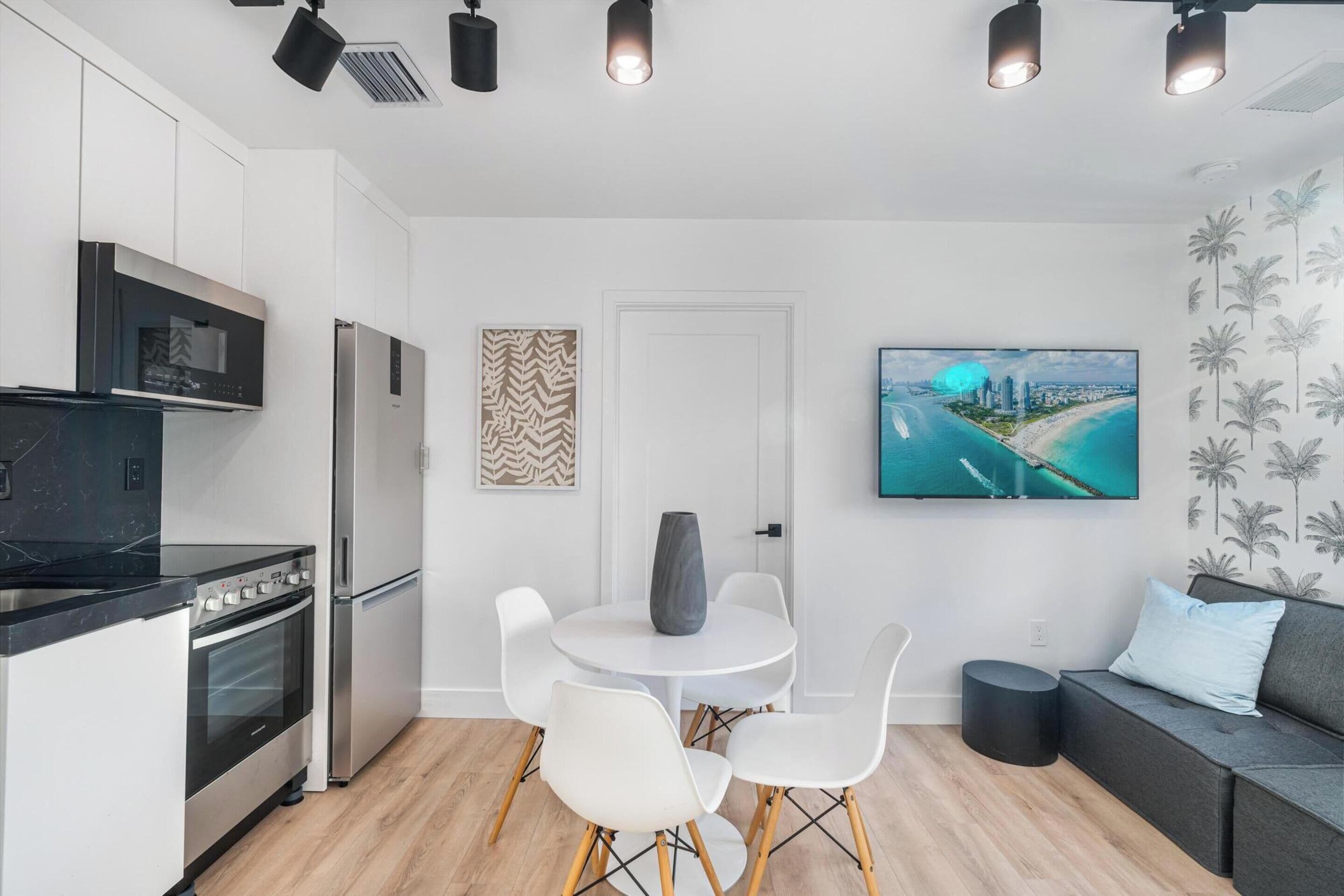 A modern kitchen with white cabinets, a black countertop, and stainless steel appliances. There is a dining area with a white table and chairs, and a black sofa with a light blue pillow. A large TV is mounted on the wall above the sofa.