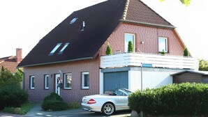 A two-story brick house with a sloped roof, a white car parked in front, and a small garden area.