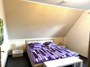 Un dormitorio con techo inclinado, una cama con colcha a rayas moradas y blancas, dos mesitas de noche con lámparas, y una pequeña mesita de noche con un adorno.