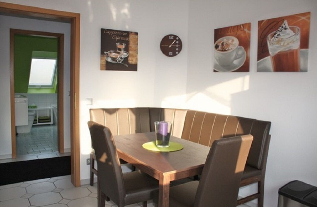 Un comedor con una mesa de madera, asientos tipo banca acolchados, un reloj y arte mural que representa bebidas.