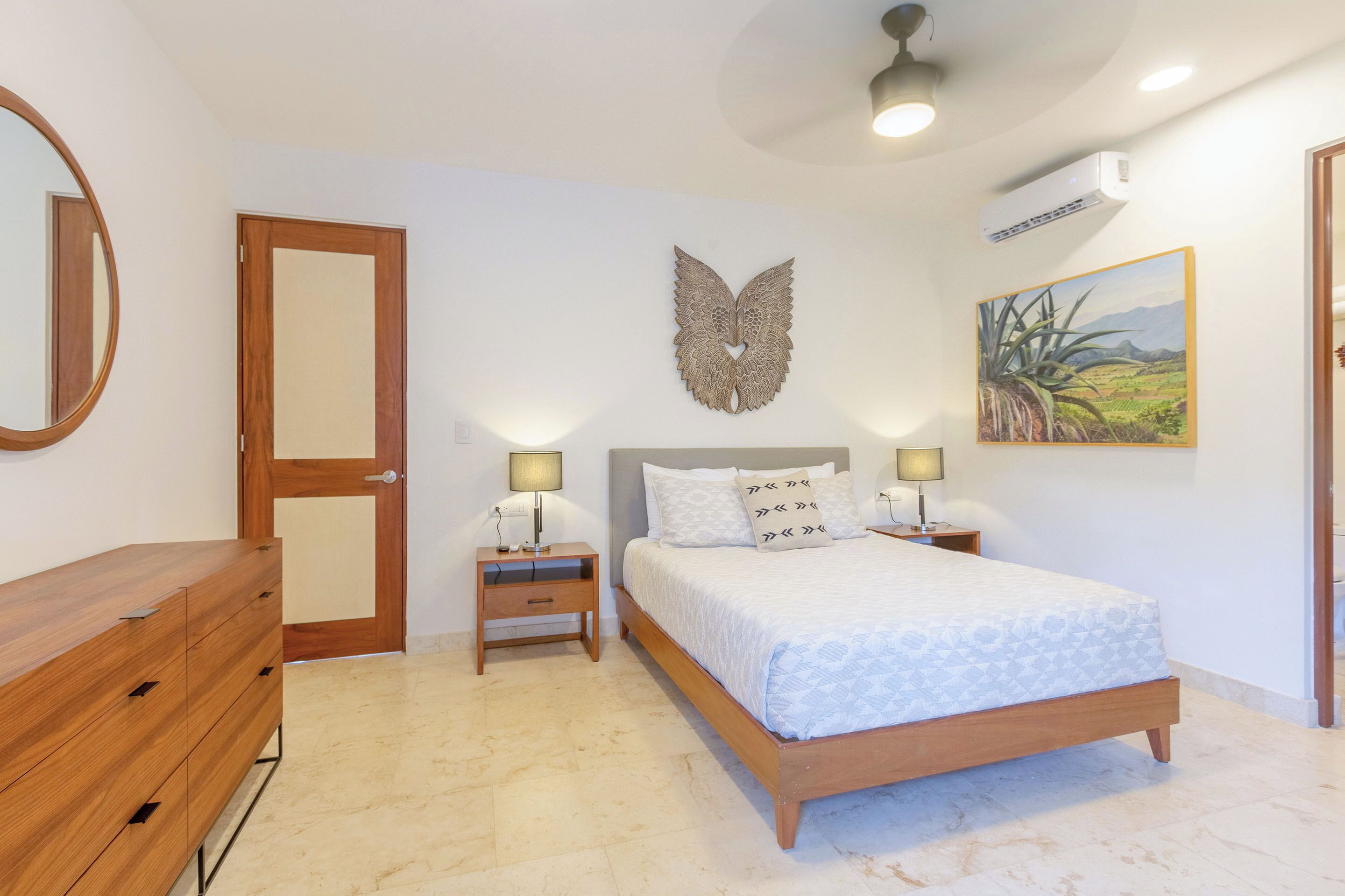 A bedroom with a wooden bed, bedside lamps, a mirror, a wall-mounted decorative piece, and a painting.