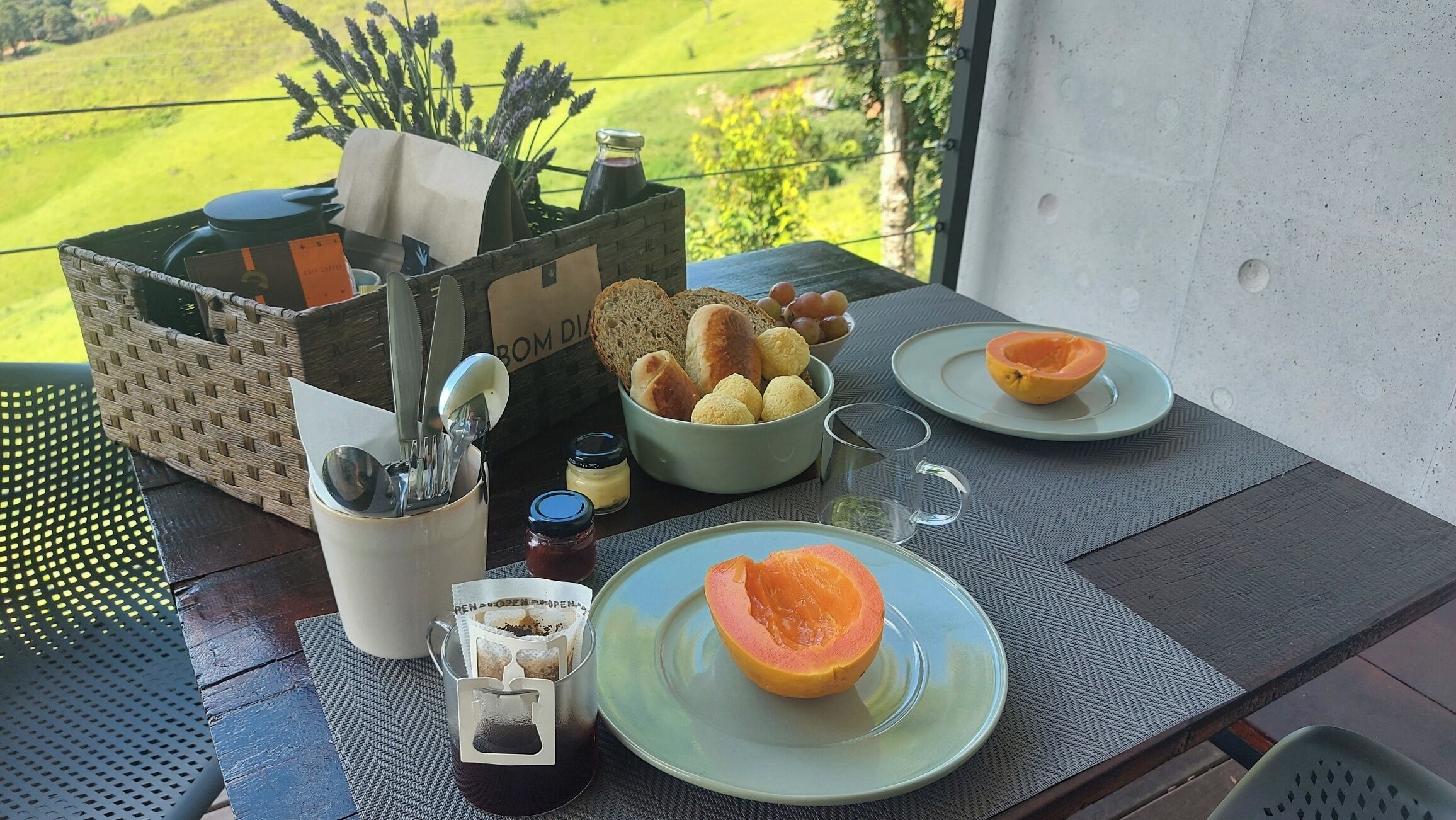 Une table dressée avec une bouteille de café, un bol de pain, une assiette avec une portion de fruit coupée en deux et un verre de jus.