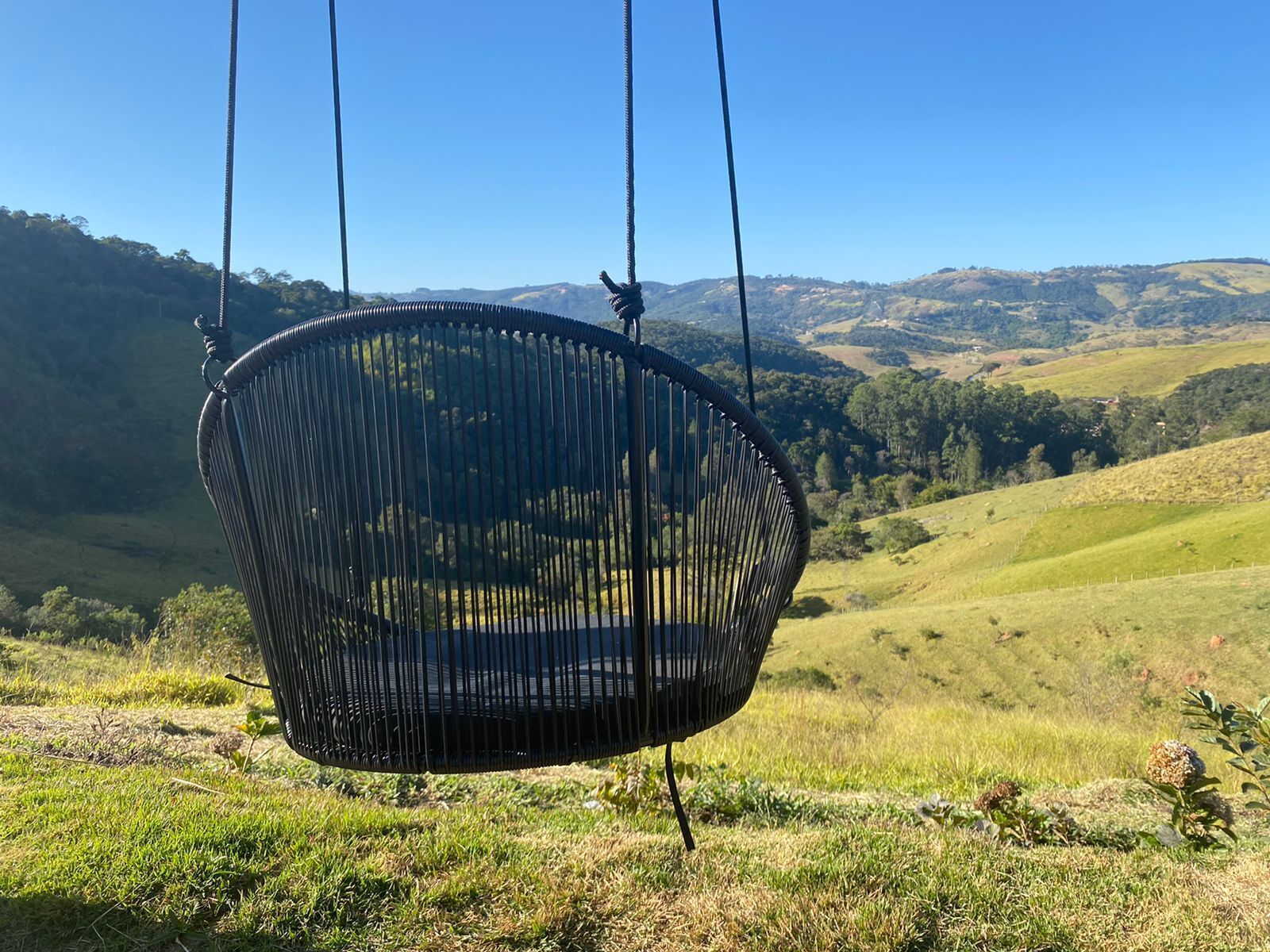 A black hanging chair suspended by ropes in a grassy field with a scenic view of rolling hills and a clear sky.