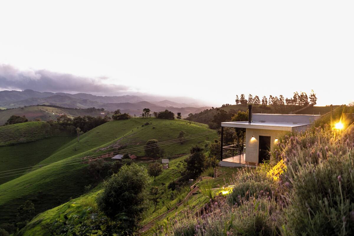 A modern house with a flat roof and a balcony, surrounded by lush green hills and a valley.