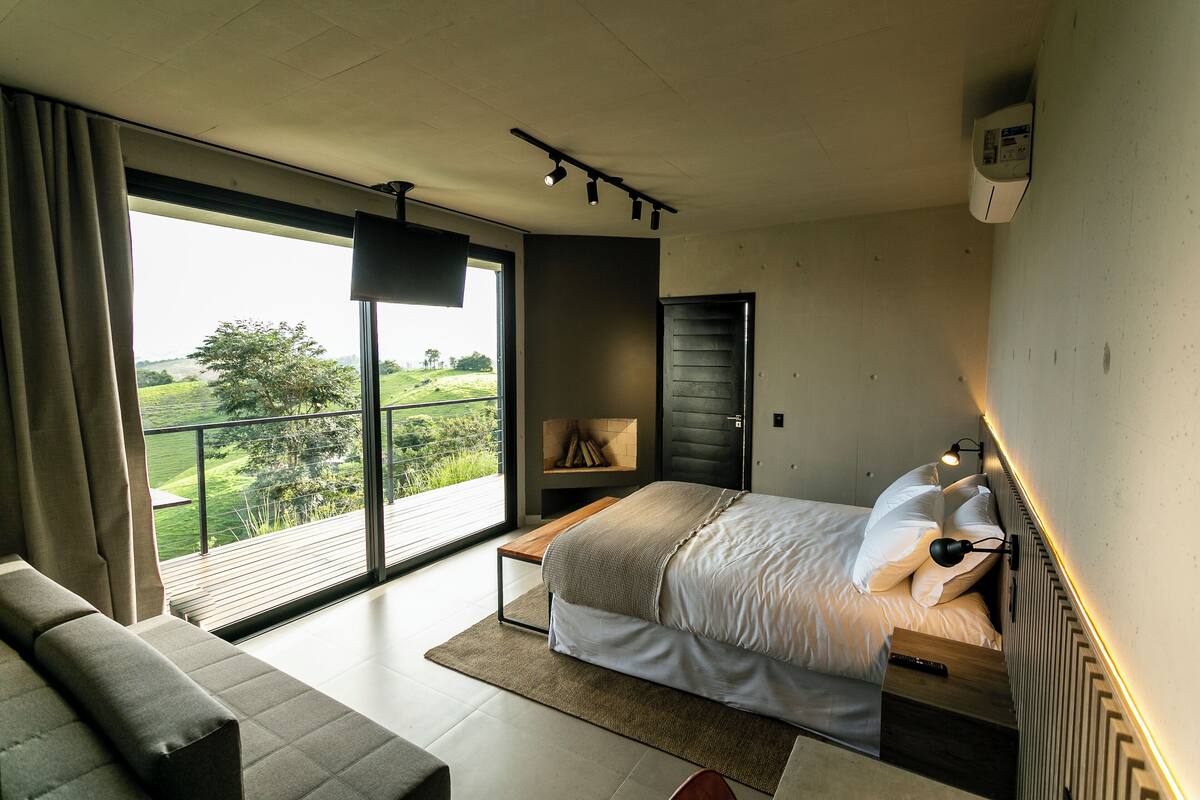A bedroom with a large window, a bed, a bench, and a wall-mounted heater.