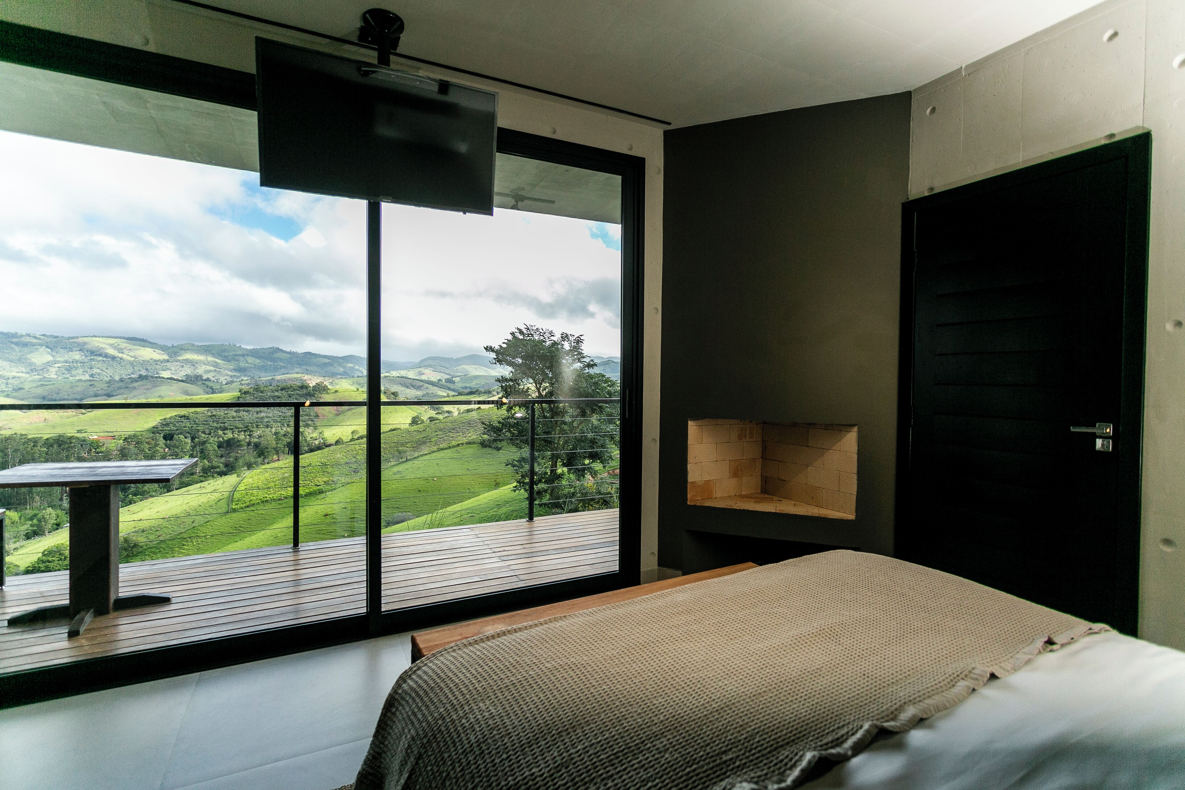 A bedroom with a large window offering a view of green hills and a cloudy sky.
