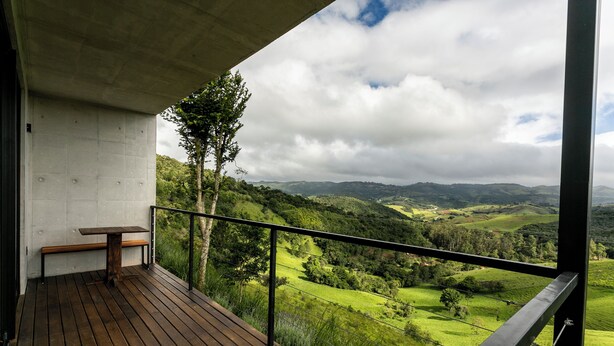 Un balcón con mesa y banco de madera, con vistas a un valle exuberante y montañas a lo lejos.