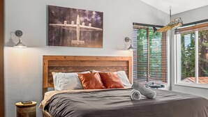 Une chambre à coucher avec une tête de lit en bois, un grand lit avec une literie sombre, deux lampes de chevet, une table de chevet avec un livre, un tableau représentant un avion et une fenêtre avec des stores.