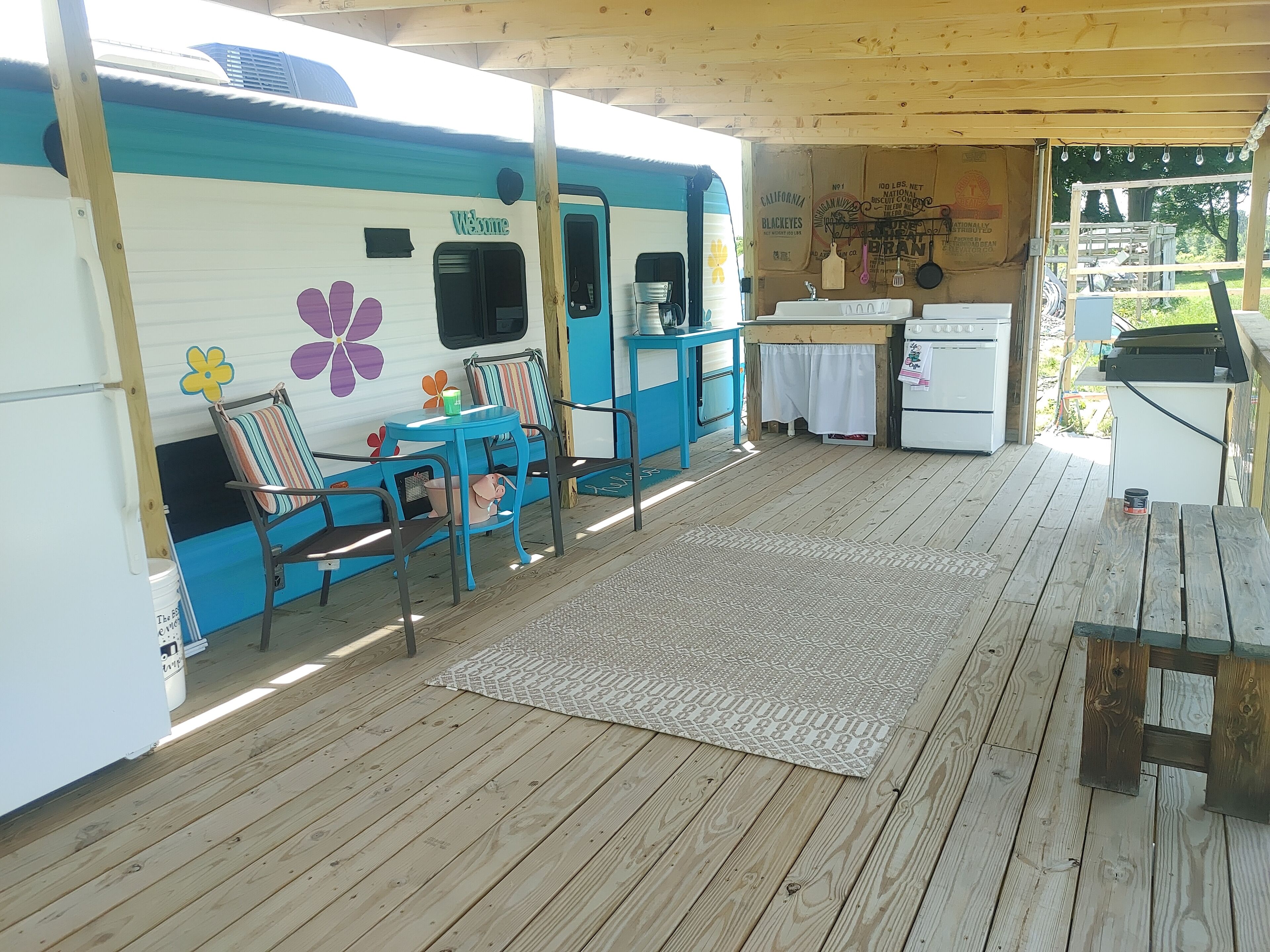 A covered outdoor area with a wooden deck, a blue and white RV, a small kitchen area, and a seating area with a table and chairs.