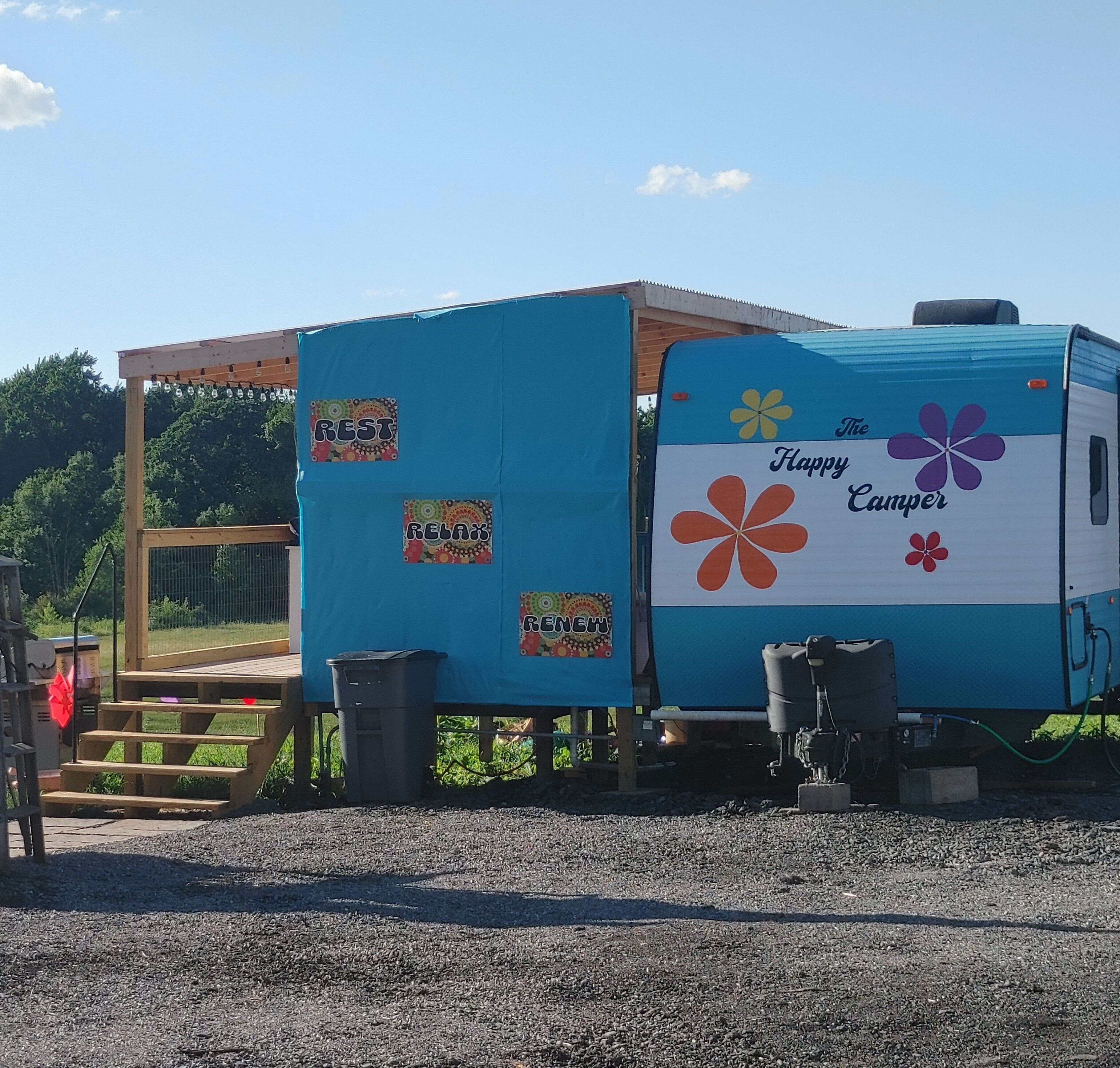 A blue camper with 'Happy Camper' and flower decals, a covered area with steps, and a gravel ground.