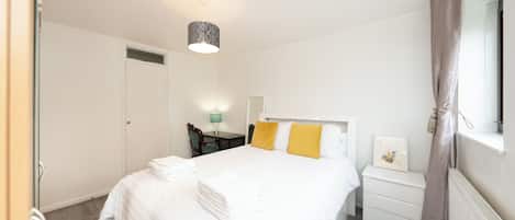 Ein Hotelzimmer mit einem ordentlich bezogenen Bett, einem Nachttisch mit Lampe, einem Sessel und einem Fenster mit Vorhängen.