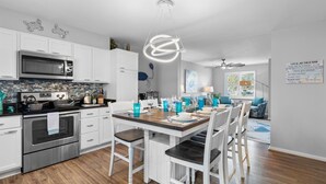 A modern kitchen with white cabinetry, a stainless steel stove, and a dining area with a wooden table and white chairs. - Central Location~Walk 2 Dining! Beach Pass & Gear! (Chincoteague)